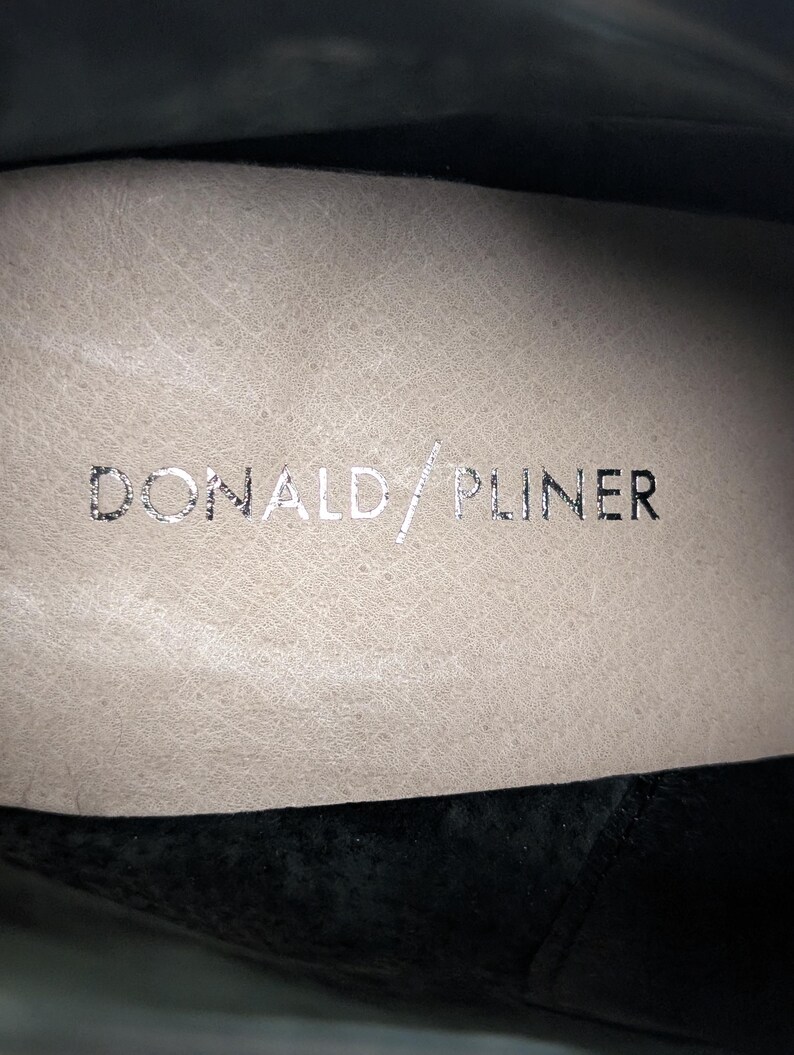 May include: Close-up of the interior of a shoe with the brand name "DONALD/PLINER" in silver lettering on a beige leather insole. The shoe's interior is visible, with a black suede exterior.