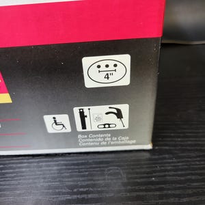 May include: A black and white illustration of a wheelchair, a faucet, and a pipe wrench. The text "Box Contents, Contenido de la Caja, Contenu de l'emballage" is printed below the illustrations.