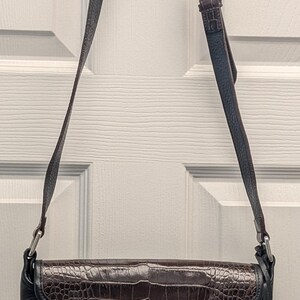 May include: Black leather crossbody bag with a brown, textured flap. The bag has a long, adjustable strap and a silver buckle. A silver heart-shaped charm hangs from the side. The bag is hanging on a white door.