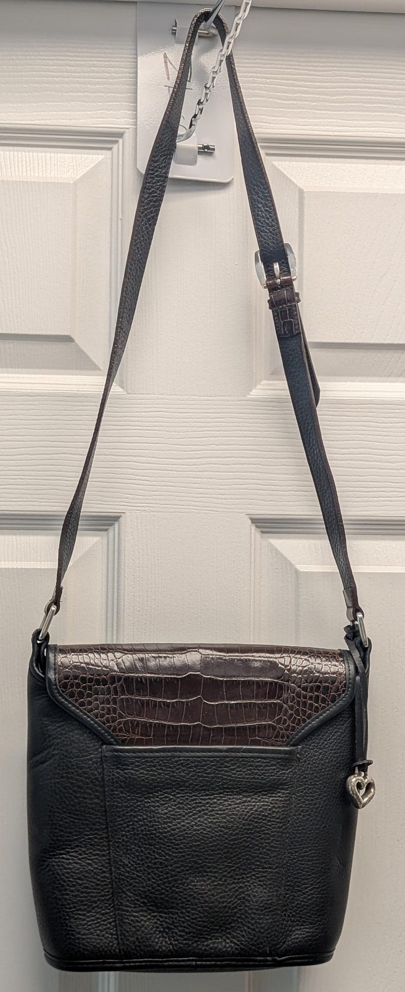 May include: Black leather crossbody bag with a brown, textured flap. The bag has a long, adjustable strap and a silver buckle. A silver heart-shaped charm hangs from the side. The bag is hanging on a white door.