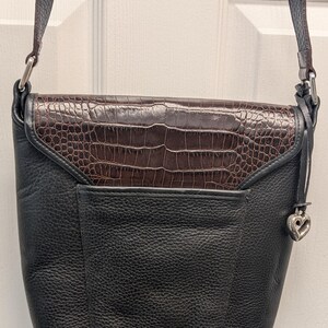 May include: Black leather crossbody bag with a brown, crocodile-textured flap. The bag has a silver-tone strap and a heart-shaped charm. The bag is designed with a front pocket and a long strap for shoulder or crossbody wear.