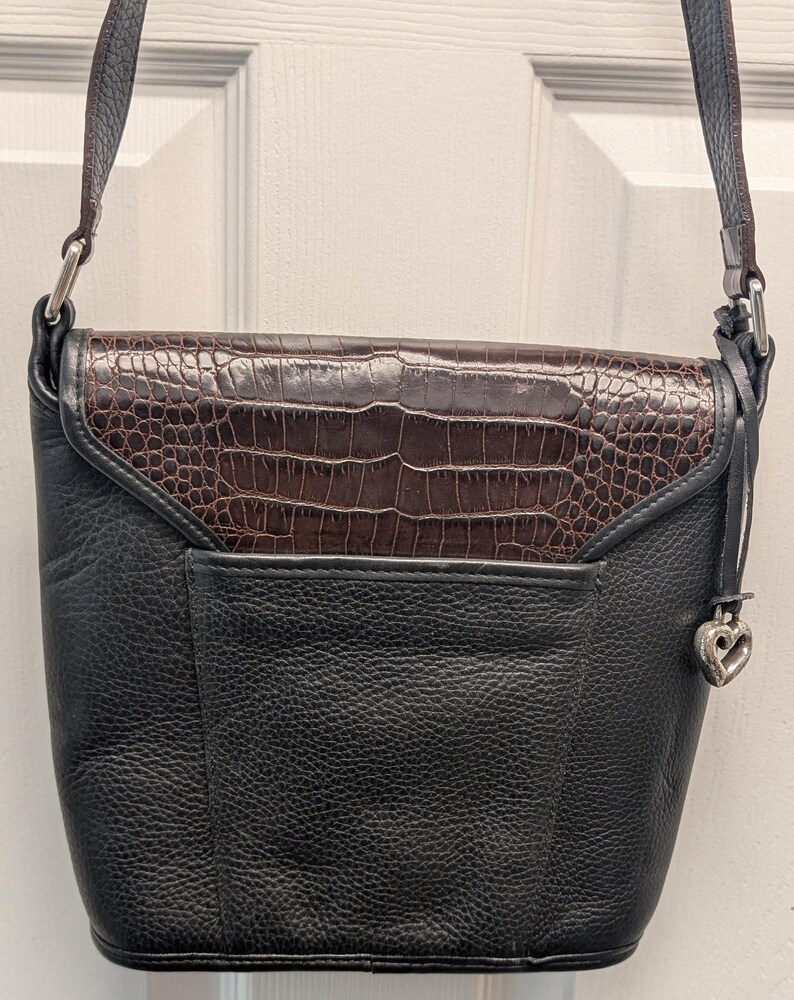 May include: Black leather crossbody bag with a brown, crocodile-textured flap. The bag has a silver-tone strap and a heart-shaped charm. The bag is designed with a front pocket and a long strap for shoulder or crossbody wear.