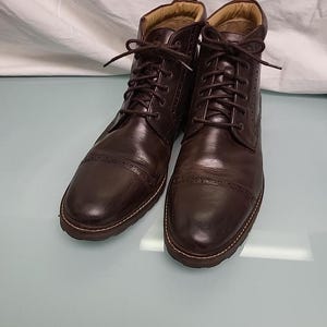 May include: A pair of dark brown leather boots with a lace-up design. The boots have a cap toe and decorative stitching. The soles are a lighter brown color. The boots are displayed on a light blue surface.