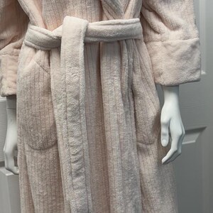 May include: A light pink, plush robe with a matching belt, displayed on a mannequin. The robe has a textured, ribbed design and long sleeves. The robe is made of a soft, comfortable material.