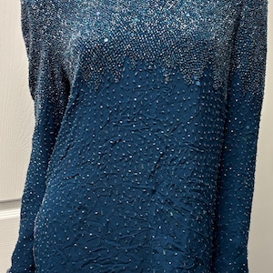 May include: Teal-colored long-sleeve top with a V-neckline. The top is heavily embellished with sequins and beads, creating a textured, shimmering effect. The sleeves and neckline are trimmed with a beaded border. The garment appears to be vintage or retro.