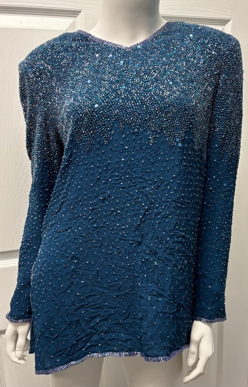 May include: Teal-colored long-sleeve top with a V-neckline. The top is heavily embellished with sequins and beads, creating a textured, shimmering effect. The sleeves and neckline are trimmed with a beaded border. The garment appears to be vintage or retro.