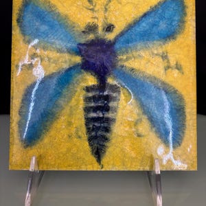 May include: Square ceramic tile with a hand-painted design of a blue and black insect on a yellow background. The insect has large, detailed wings. The tile is displayed on a clear acrylic stand.