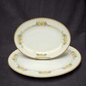May include: Two oval serving platters with a cream-colored base. Each platter features a gold rim and a floral design in yellow, pink, and blue. The platters are stacked on top of each other, showcasing their decorative details.