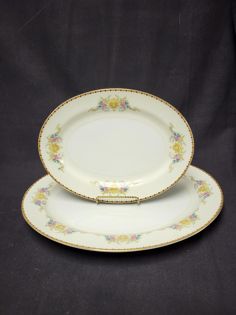 May include: Two oval serving platters with a cream-colored base. Each platter features a gold rim and a floral design in yellow, pink, and blue. The platters are stacked on top of each other, showcasing their decorative details.