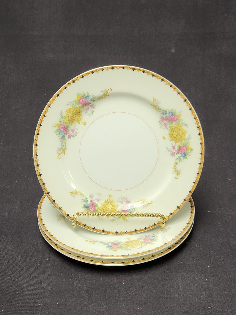 May include: A stack of three white porcelain plates with a gold rim and floral design. The plates have a gold and black star pattern around the edge and a floral design with pink, yellow, and blue flowers.
