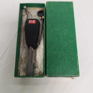 May include: A vintage black microphone with the letters "K-E" on the front, presented in an open green box. The microphone has a textured surface and a gray handle. The box is lined with tissue paper.