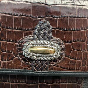 May include: Close-up of a brown leather handbag with a crocodile skin texture. The bag features a silver and gold-toned clasp with a braided leather accent. The bag's design includes a black trim.