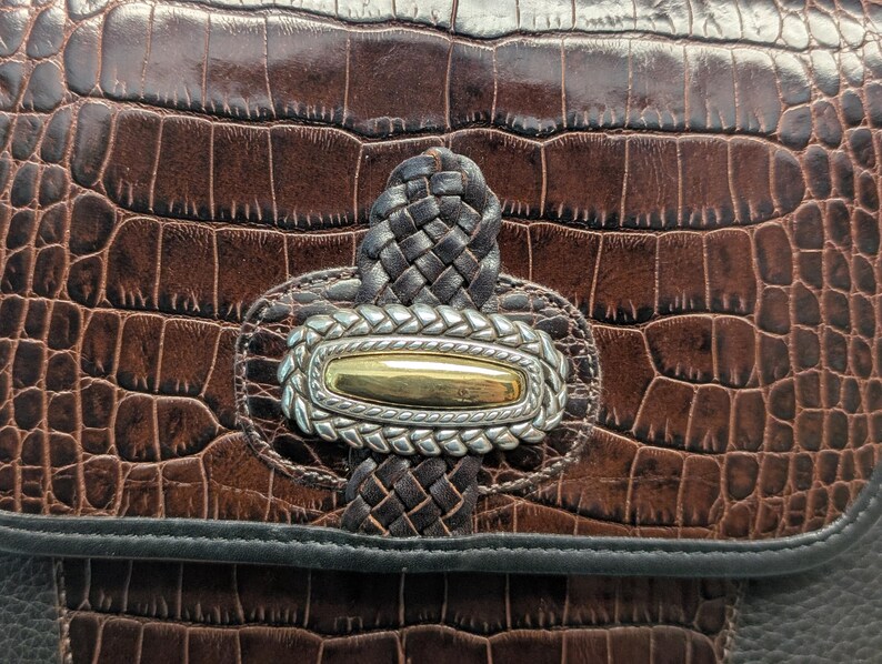 May include: Close-up of a brown leather handbag with a crocodile skin texture. The bag features a silver and gold-toned clasp with a braided leather accent. The bag's design includes a black trim.