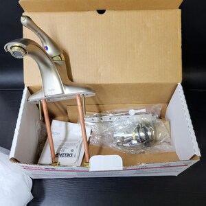 May include: A new, unopened Delta brand bathroom faucet in a brushed nickel finish, in its original box. The faucet is a two-handle design with a spout and two handles.
