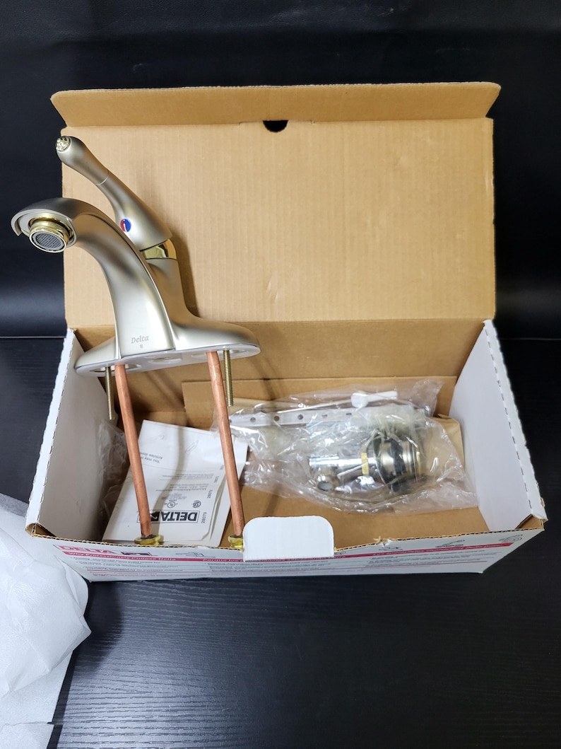 May include: A new, unopened Delta brand bathroom faucet in a brushed nickel finish, in its original box. The faucet is a two-handle design with a spout and two handles.