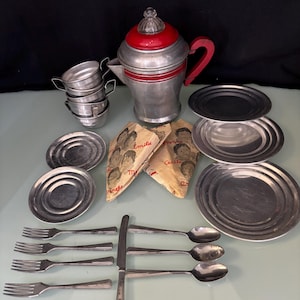 May include: A vintage toy tea set with a silver finish, including a teapot with a red handle and lid, cups, saucers, plates, forks, spoons, and a knife. Two small fabric packets with illustrations are also included.