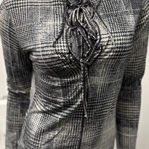 May include: A black and white houndstooth print top with a high neckline and long sleeves. The top features a decorative lace-up detail at the chest. The fabric has a subtle sheen, and the overall style is classic and versatile.