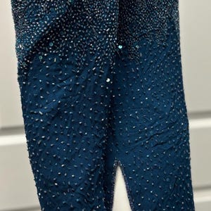 May include: Teal-colored beaded jacket with long sleeves and a split up the side. The top portion of the jacket is densely covered in beads, transitioning to a sparser pattern towards the bottom. The edges are trimmed with a lighter blue.