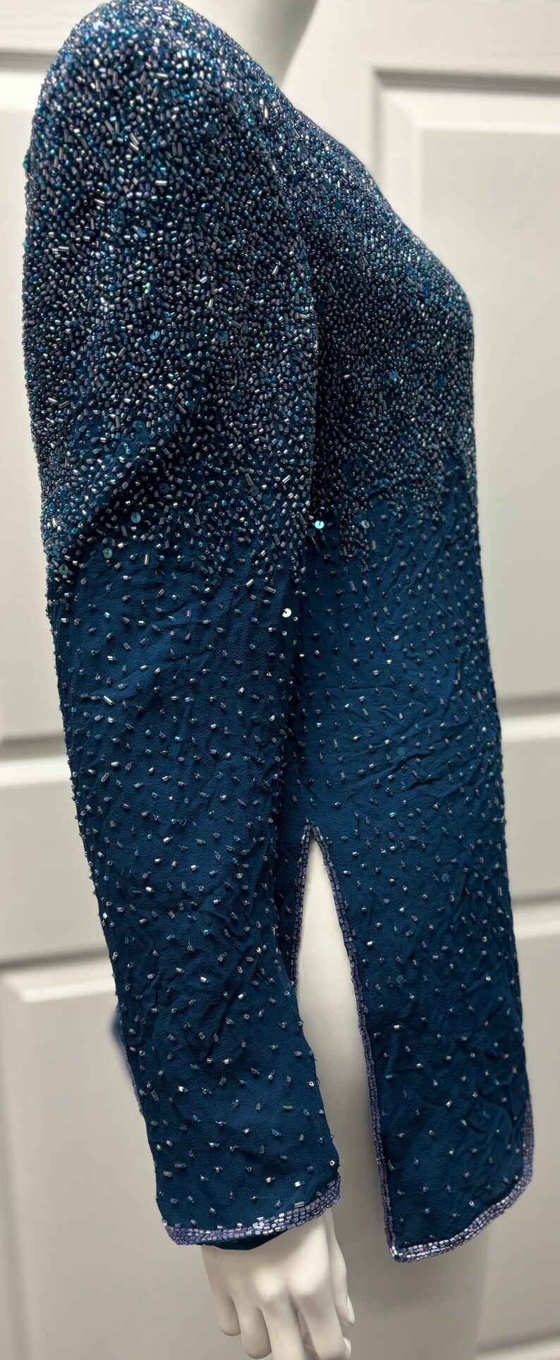May include: Teal-colored beaded jacket with long sleeves and a split up the side. The top portion of the jacket is densely covered in beads, transitioning to a sparser pattern towards the bottom. The edges are trimmed with a lighter blue.
