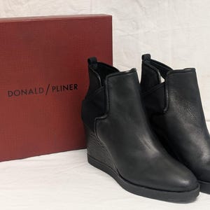 May include: A pair of black leather wedge ankle boots with a side elastic panel. The boots are displayed next to a red box with the brand name "DONALD / PLINER" printed in white.