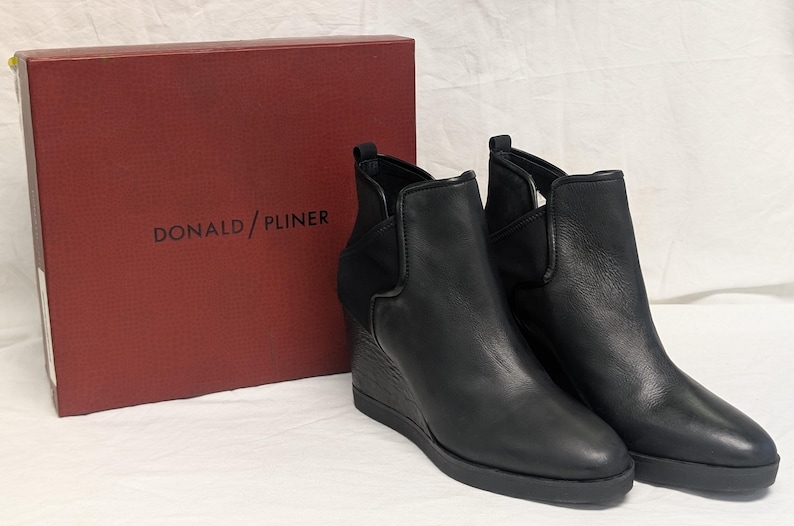 May include: A pair of black leather wedge ankle boots with a side elastic panel. The boots are displayed next to a red box with the brand name "DONALD / PLINER" printed in white.