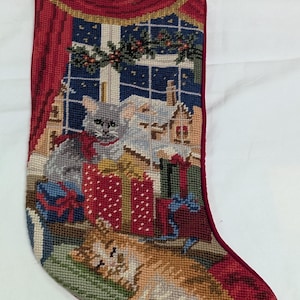 May include: A needlepoint Christmas stocking featuring a festive scene with a gray cat sitting near wrapped gifts and a ginger cat lying on a green rug. The background shows a window with red curtains, a snowy landscape, and a holiday garland.