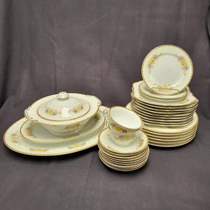 May include: A complete vintage porcelain dinnerware set. The set includes a covered serving dish, oval platter, plates, saucers, and teacups. Each piece is white with a floral design in yellow, pink, and blue, and gold trim.