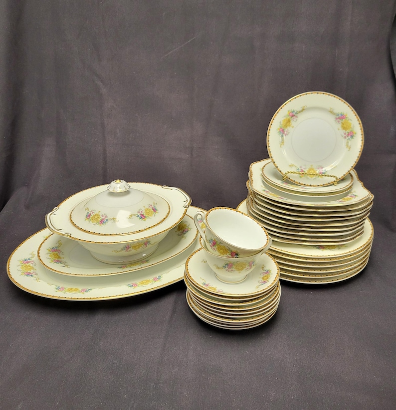 May include: A complete vintage porcelain dinnerware set. The set includes a covered serving dish, oval platter, plates, saucers, and teacups. Each piece is white with a floral design in yellow, pink, and blue, and gold trim.