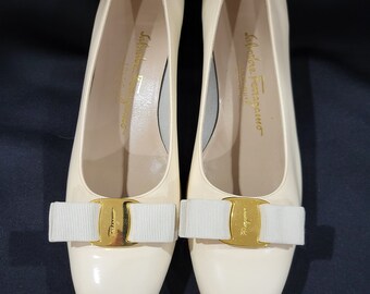 May include: A pair of cream-colored high heels with a glossy finish. Each shoe features a white ribbon and a gold-colored buckle. The brand name "Salvatore Ferragamo" is printed in gold on the insole.