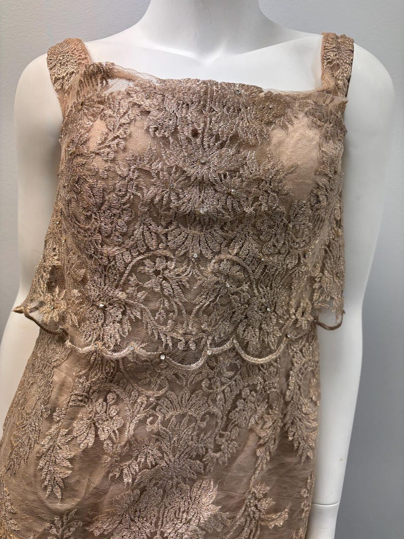 Pode incluir: Um vestido sem mangas feito de renda bege delicada com um padr&atilde;o floral. O vestido tem um decote quadrado e uma saia em camadas.