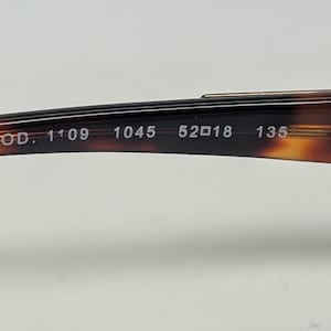 May include: Close-up of the arm of a pair of eyeglasses with a tortoiseshell pattern. The text "MOD. 1'09 1045 52018 135" is printed on the arm. The glasses have a curved design and are made of a glossy material.