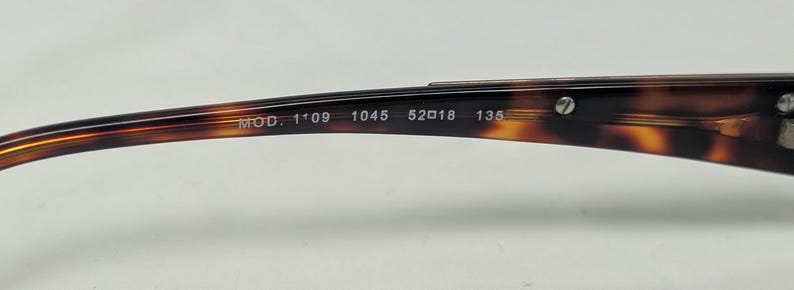 May include: Close-up of the arm of a pair of eyeglasses with a tortoiseshell pattern. The text "MOD. 1'09 1045 52018 135" is printed on the arm. The glasses have a curved design and are made of a glossy material.