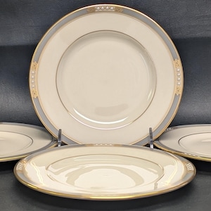 May include: A set of four white porcelain plates with a gold rim and a gray border. The plates are arranged on a dark surface, with one plate on a stand. The plates have a simple, elegant design.
