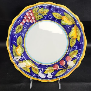May include: A round, decorative ceramic plate with a white center and a blue border. The border is adorned with a yellow scalloped edge and a colorful fruit design featuring grapes, pears, and cherries. The plate is displayed on a clear stand.
