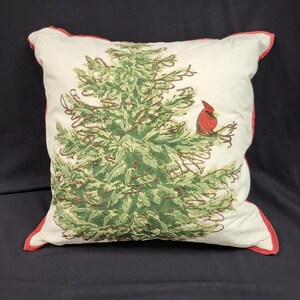 May include: Square decorative pillow with a cream-colored background. The pillow features a detailed green Christmas tree design with a red cardinal bird perched on a branch. The edges are trimmed with a red border.