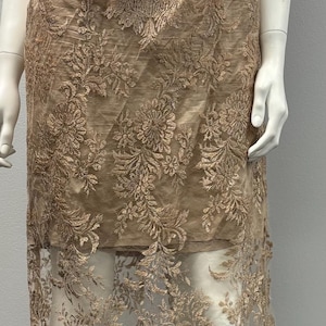 Pode incluir: Um vestido comprido sem mangas com uma sobreposi&ccedil;&atilde;o transparente. O vestido &eacute; feito de um delicado tecido de renda floral em uma cor marrom claro.