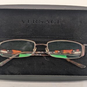 May include: Rectangular eyeglasses with a brown frame and tortoise shell accents, resting in an open black case. The case is embossed with the brand name "VERSACE". The glasses have a metal frame and clear lenses.