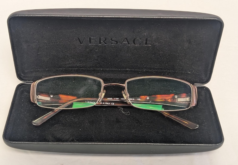 May include: Rectangular eyeglasses with a brown frame and tortoise shell accents, resting in an open black case. The case is embossed with the brand name "VERSACE". The glasses have a metal frame and clear lenses.