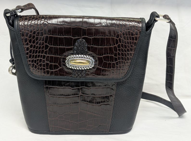 May include: A black and brown crossbody bag with a textured, faux crocodile pattern. The bag features a flap closure with a decorative silver and gold accent. The adjustable strap is black, and the bag has a structured shape.
