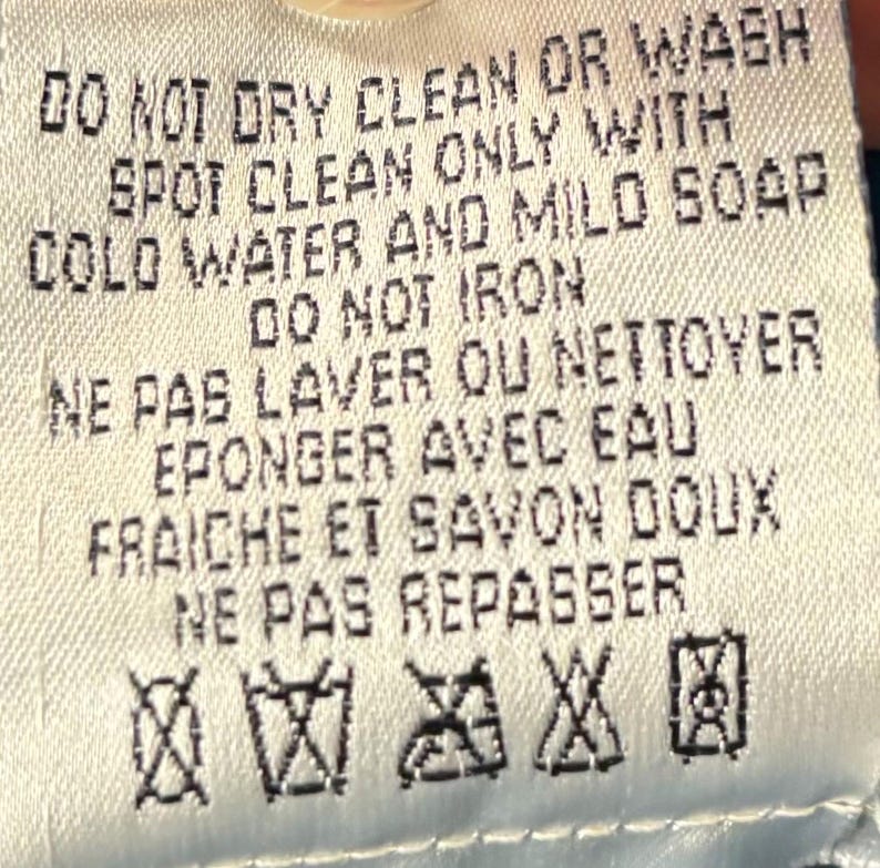 May include: A close-up of a white fabric tag with black text. The tag provides care instructions: "DO NOT DRY CLEAN OR WASH", "SPOT CLEAN ONLY", "DO NOT IRON", and other washing instructions. There are also several laundry symbols.