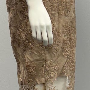 Pode incluir: Um vestido longo sem mangas com um delicado padr&atilde;o de renda floral em cor marrom claro. O vestido tem uma sobreposi&ccedil;&atilde;o transparente com um padr&atilde;o de renda semelhante.