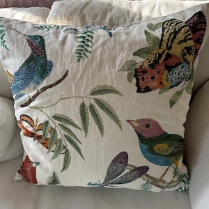 May include: A decorative throw pillow with a white background featuring colorful illustrations of birds, butterflies, and dragonflies. The pillow has a botanical theme with detailed drawings of leaves and branches.