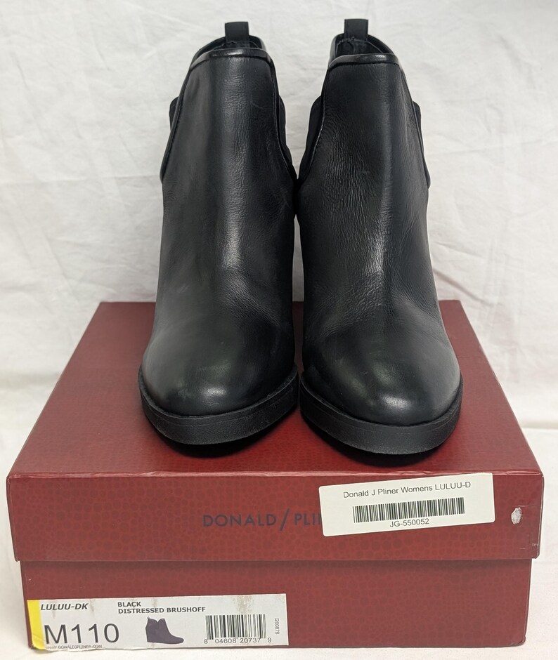 May include: A pair of black leather ankle boots with a rounded toe and a stacked heel. The boots are displayed on a red box with the brand name "DONALD/PLINER" visible. The box also has product information including the style name "LULUU-DK".