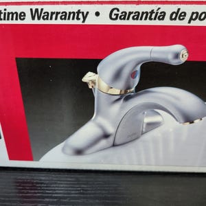 May include: A silver faucet with a gold handle and a white background. The faucet has a single handle and a spout. The text "Lifetime Warranty" and "Garant&iacute;a de por vida" is visible in the background.