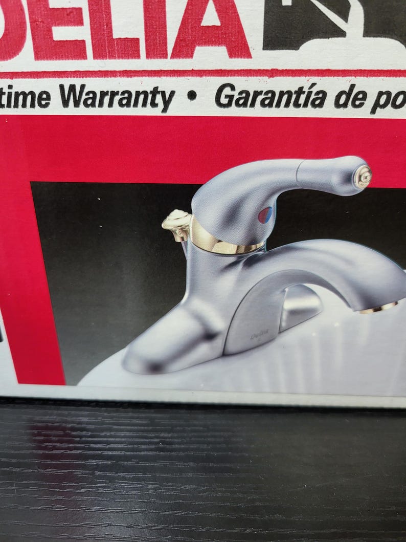 May include: A silver faucet with a gold handle and a white background. The faucet has a single handle and a spout. The text "Lifetime Warranty" and "Garant&iacute;a de por vida" is visible in the background.