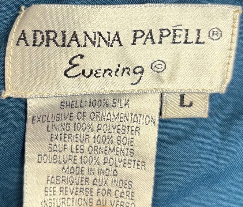 May include: Close-up of a label from an Adrianna Papell Evening garment. The label is cream-colored with black text, including the brand name, "Evening," and fabric composition details. The size is indicated as "L."