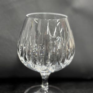 May include: A clear, stemmed brandy snifter with a wide bowl and a short stem. The glass has vertical, textured lines that run from the base to the rim. The glass sits on a round base.