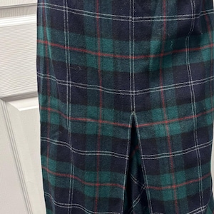May include: A knee-length plaid skirt in shades of green, navy, red, and white. The skirt features a classic tartan pattern and a front slit. The fabric appears to be a medium-weight material.