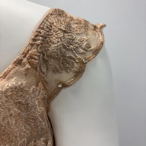Pode incluir: Close-up de um tecido de renda dourado com uma borda festoneada e pequenas p&eacute;rolas. O tecido &eacute; transparente e delicado, com um padr&atilde;o floral.