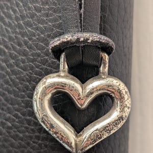 May include: Close-up of a silver heart-shaped charm with the word "Brighton" engraved on it. The charm is attached to a black leather strap, set against a textured black leather background. The charm has a distressed, vintage appearance.